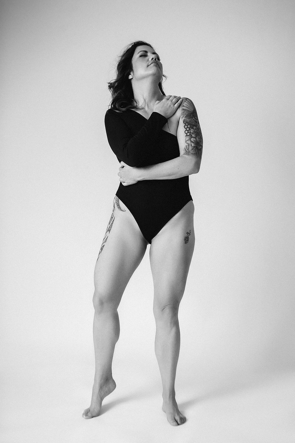A glamourous young brunette woman wears an asymmetrical bodysuit, tattoos visible. She stands tall and embraces herself