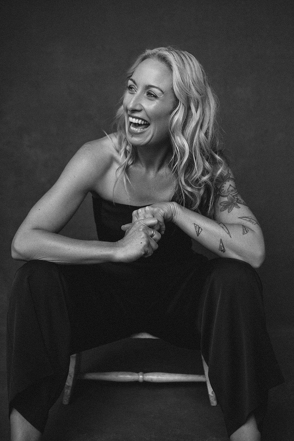 A quirky woman with long blonde hair poses on a chair. She is leaning forward and laughs broadly at something off camera