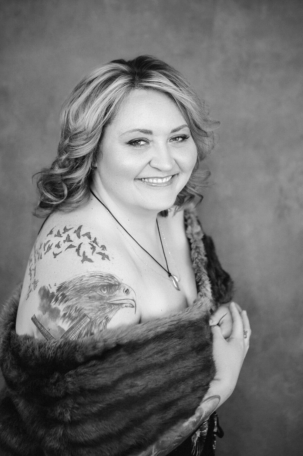 A curvy blonde woman with tattoos is wrapped in a fur, her shoulders visible as she smiles coyly at the camera