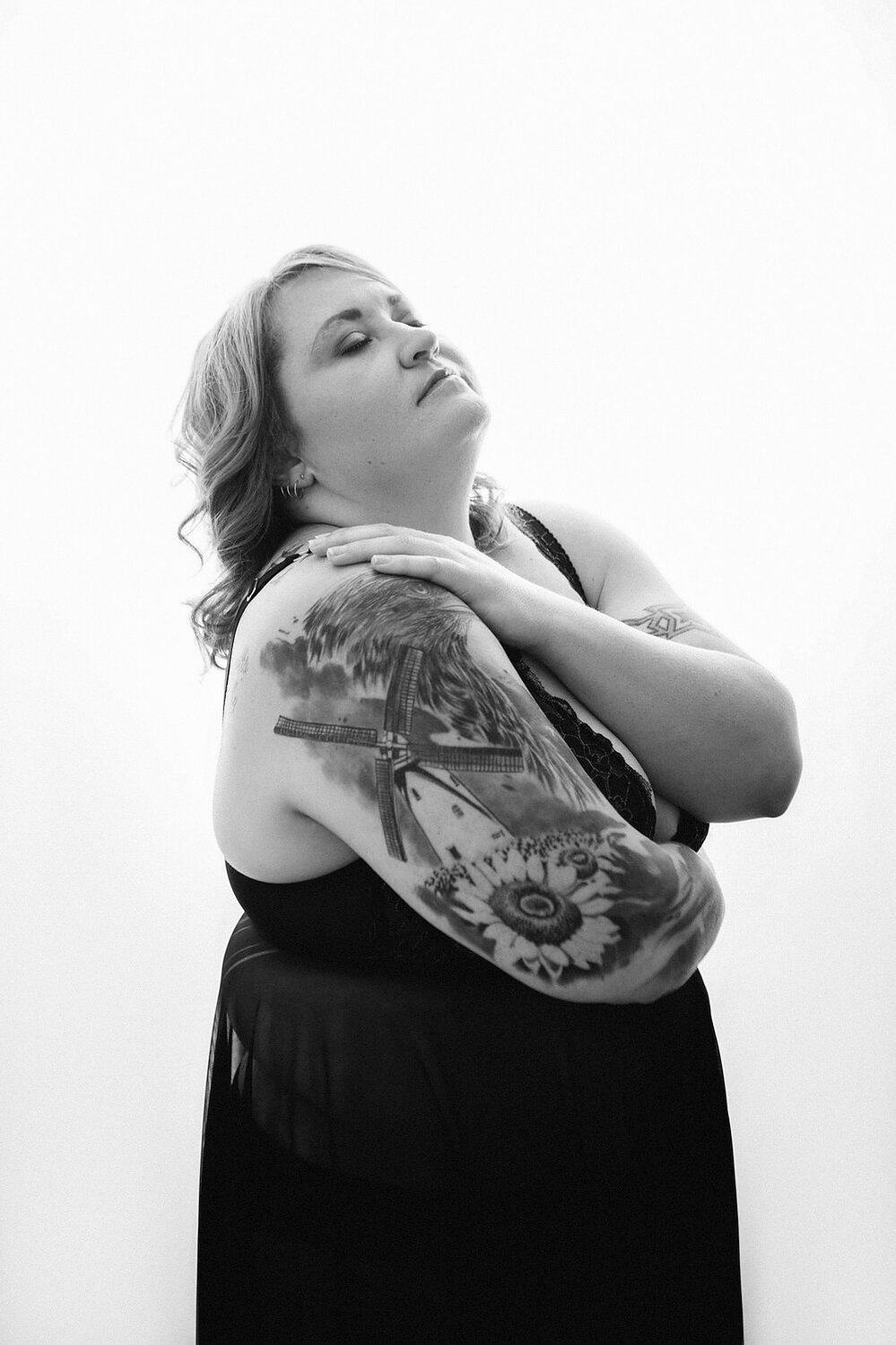 A curvy blonde woman with tattoos wears a black dress and embraces herself, eyes closed