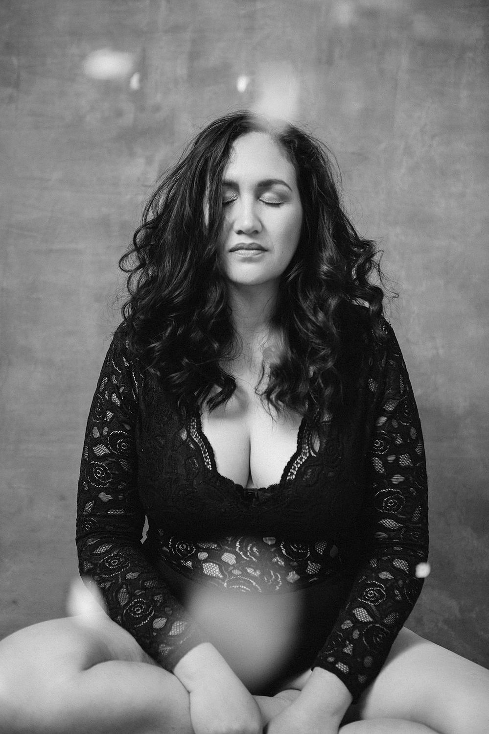 A stunning brunette woman wears a lacey bodysuit and sits with her eyes closed. Petals are falling all around her.