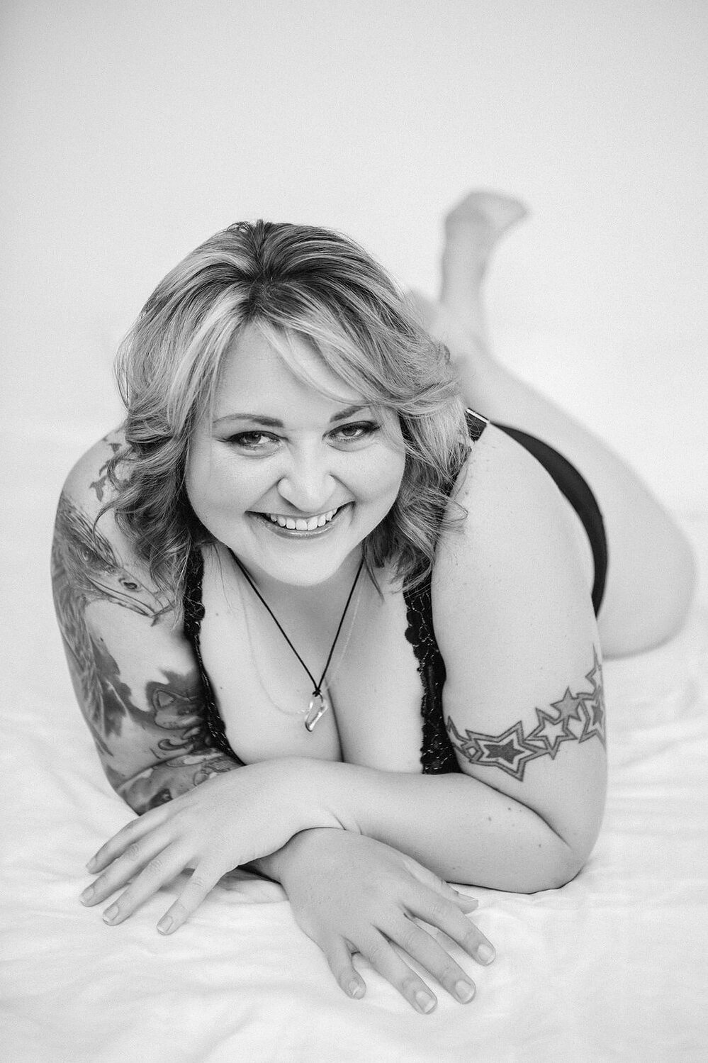 A curvy blonde woman with tattoos poses in a boudoir style, she wears lingerie and smiles at the camera