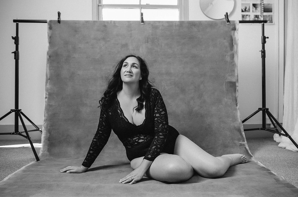 A stunning brunette woman wears a lacey bodysuit and sits in front of a photography backdrop