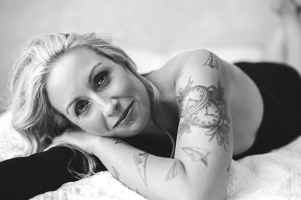 A quirky woman with long blonde hair reclines on a bed, her head resting on an arm