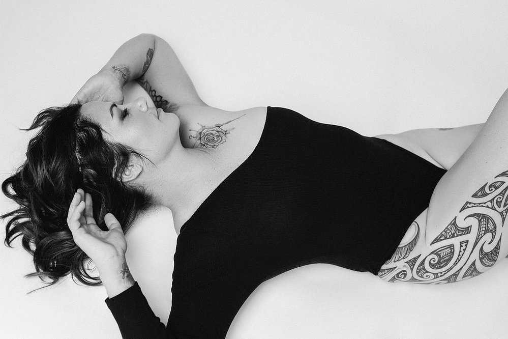 A glamourous young brunette woman wears an asymmetrical bodysuit, tattoos visible
