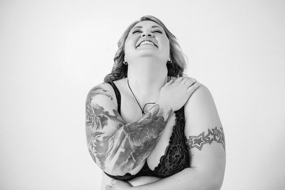 A curvy blonde woman with tattoos wears a bralette, she looks upwards as she embraces herself