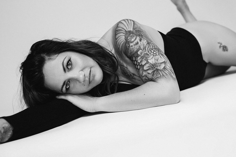 A glamourous young brunette woman wears an asymmetrical bodysuit, tattoos visible. She lies down with her head resting on an arm, gazing enigmatically at the camera