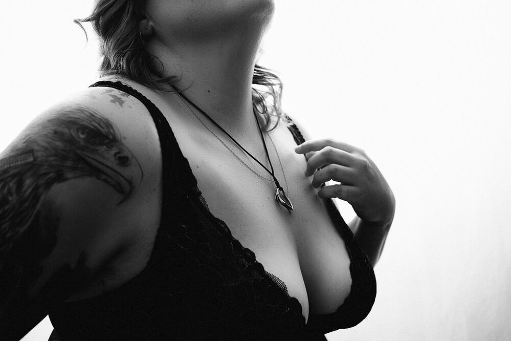 A curvy blonde woman with tattoos wears a black bralette and traces along her collarbone