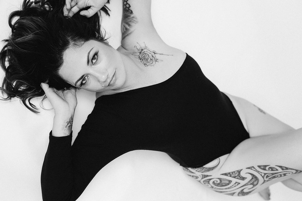A glamourous young brunette woman wears an asymmetrical bodysuit, she is reclined on the floor with hands in her hair