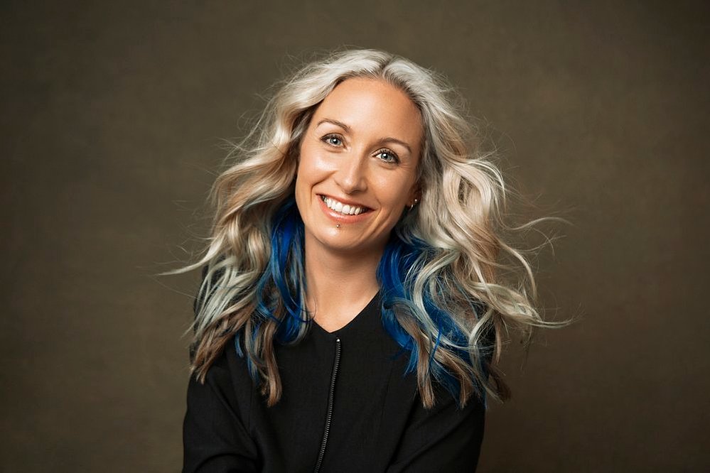 A quirky woman with long blonde hair that has blue streaks in it, she smiles enigmatically at the camera