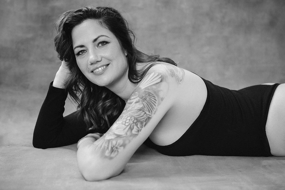 A glamourous young brunette woman wears an asymmetrical bodysuit, tattoos visible. She lies down with one hand supporting her head