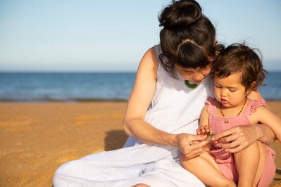 A pregnant woman with long brunette hair tied in a bun shows her toddler a shell, as they sit on a sunny golden sand beach