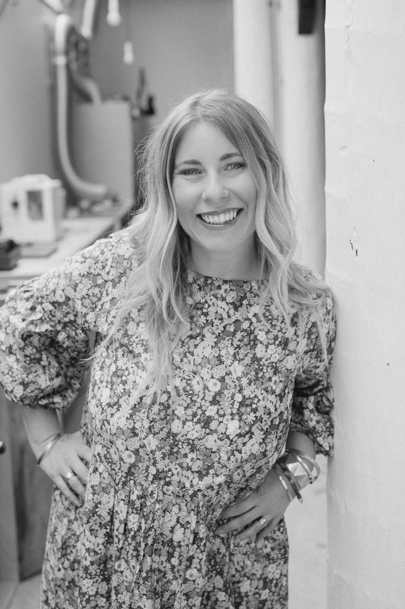 A slim blonde woman in a floral dress leans against a doorway with her hands on her hips