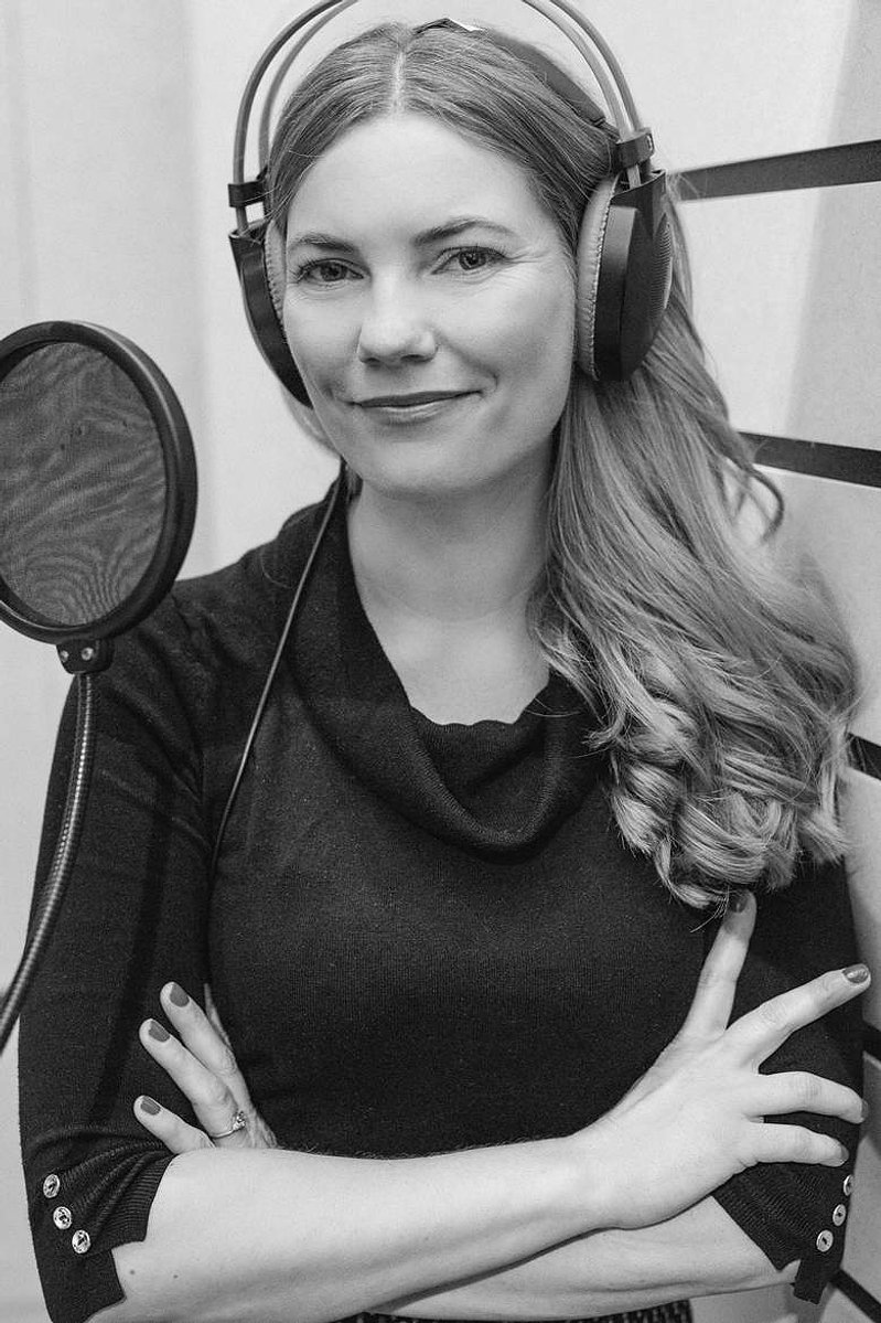 A woman with long hair stands in a recording booth, her arms a crossed in front of her and she wears headphones