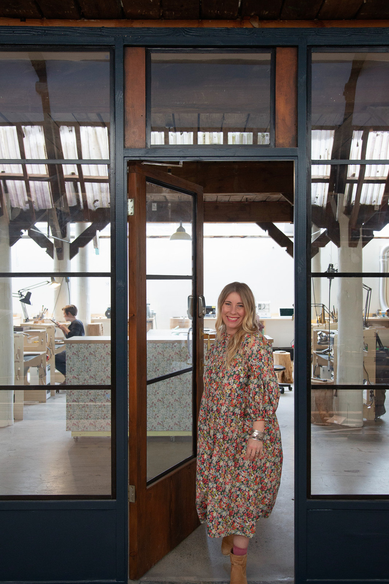 A slim blonde woman in a floral dress stands in the doorway to the jewellery workshop she works in
