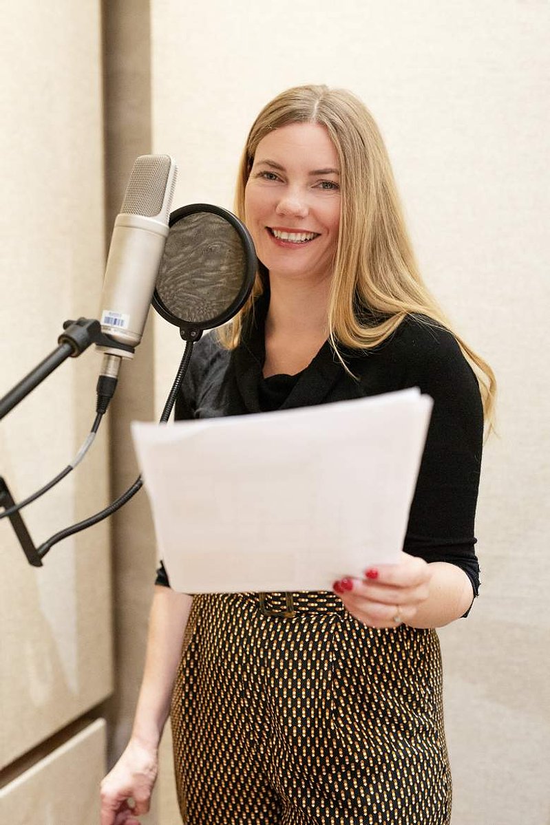 A blonde woman stands at a microphone in a recording booth, she holds a script in front of her and smiles at the camera