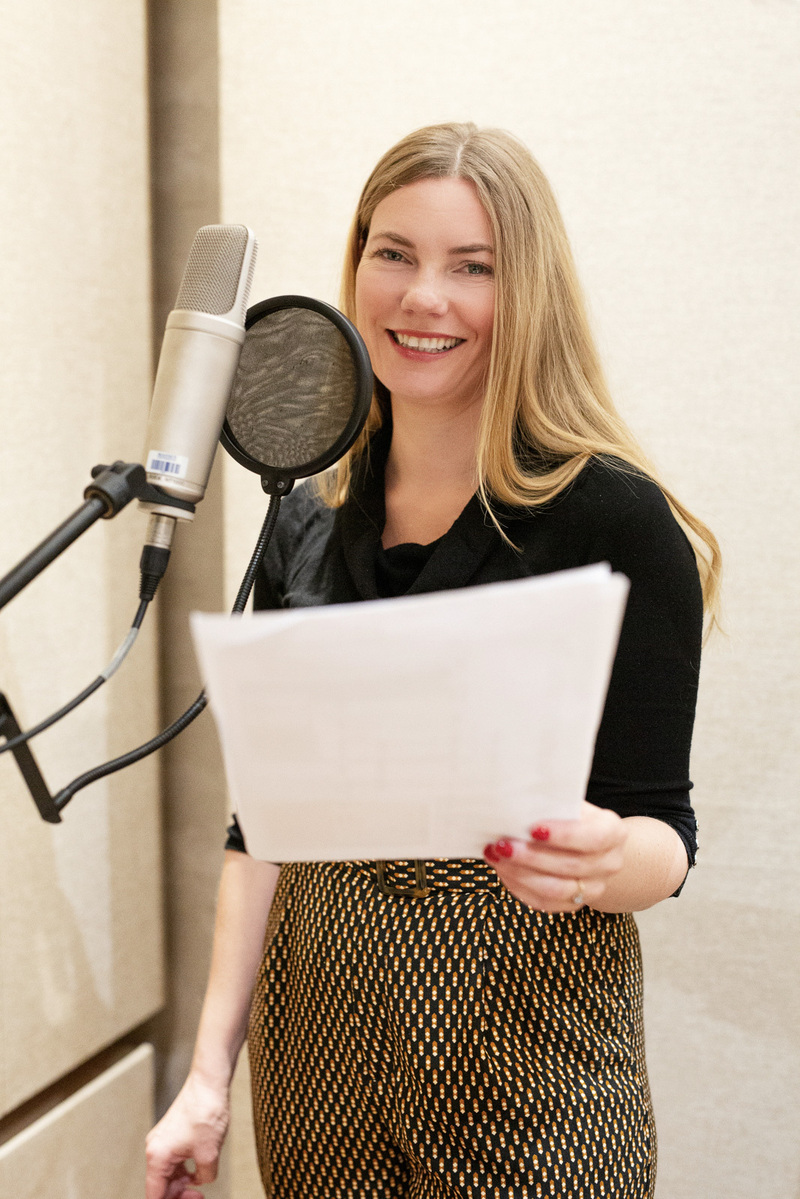 A blonde woman stands at a microphone in a recording booth, she holds a script in front of her and smiles at the camera