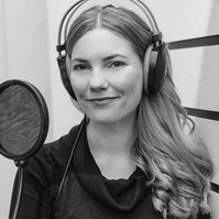 A woman with long hair stands in a recording booth, her arms a crossed in front of her and she wears headphones