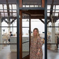 A slim blonde woman in a floral dress stands in the doorway to the jewellery workshop she works in