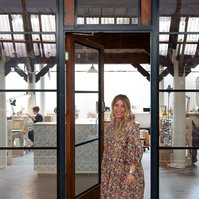 A slim blonde woman in a floral dress stands in the doorway to the jewellery workshop she works in