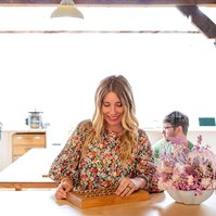 A blonde woman smiles contentedly as she looks down at a tray of handmade rings