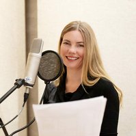 A blonde woman stands at a microphone in a recording booth, she holds a script in front of her and smiles at the camera