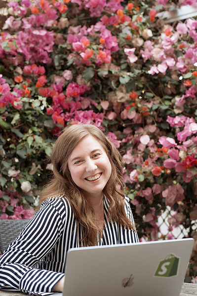 A smiling young woman sits at her laptop outside, a wall of pink flowers behind her