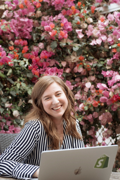 A smiling young woman sits at her laptop outside, a wall of pink flowers behind her