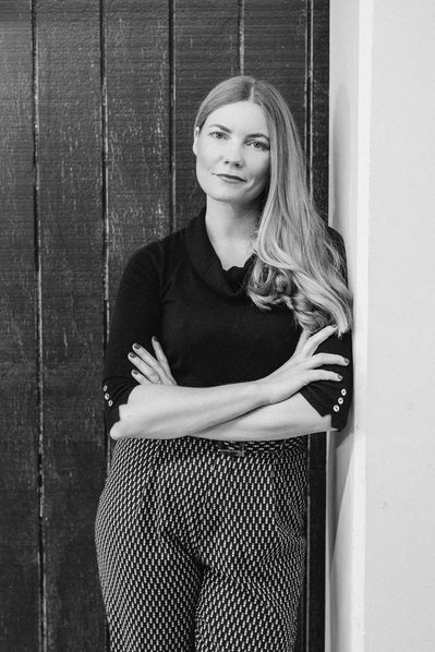 A slim woman leans against a doorway with her arms crossed, looking directly at the camera