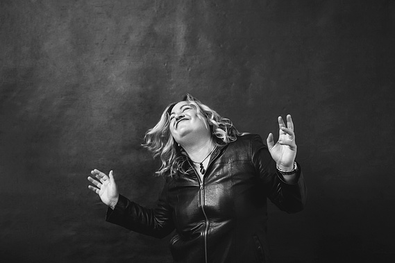 A curvaceous blonde woman dances in celebration, she is wearing a leather jacket and her hair is shaken free