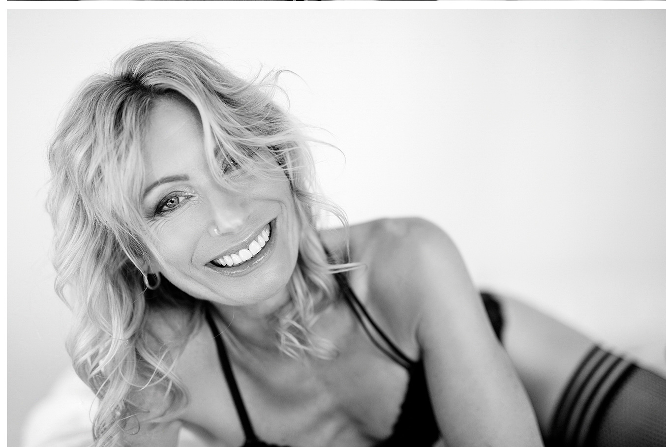 A beautiful blonde woman smiles widely at the camera as she reclines on a bed