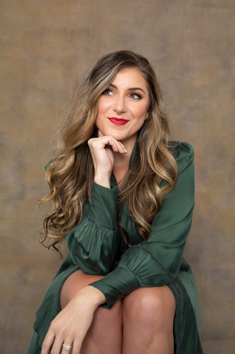 A young woman with long curled hair sits with her chin resting on a hand, she smiles enigmatically off camera
