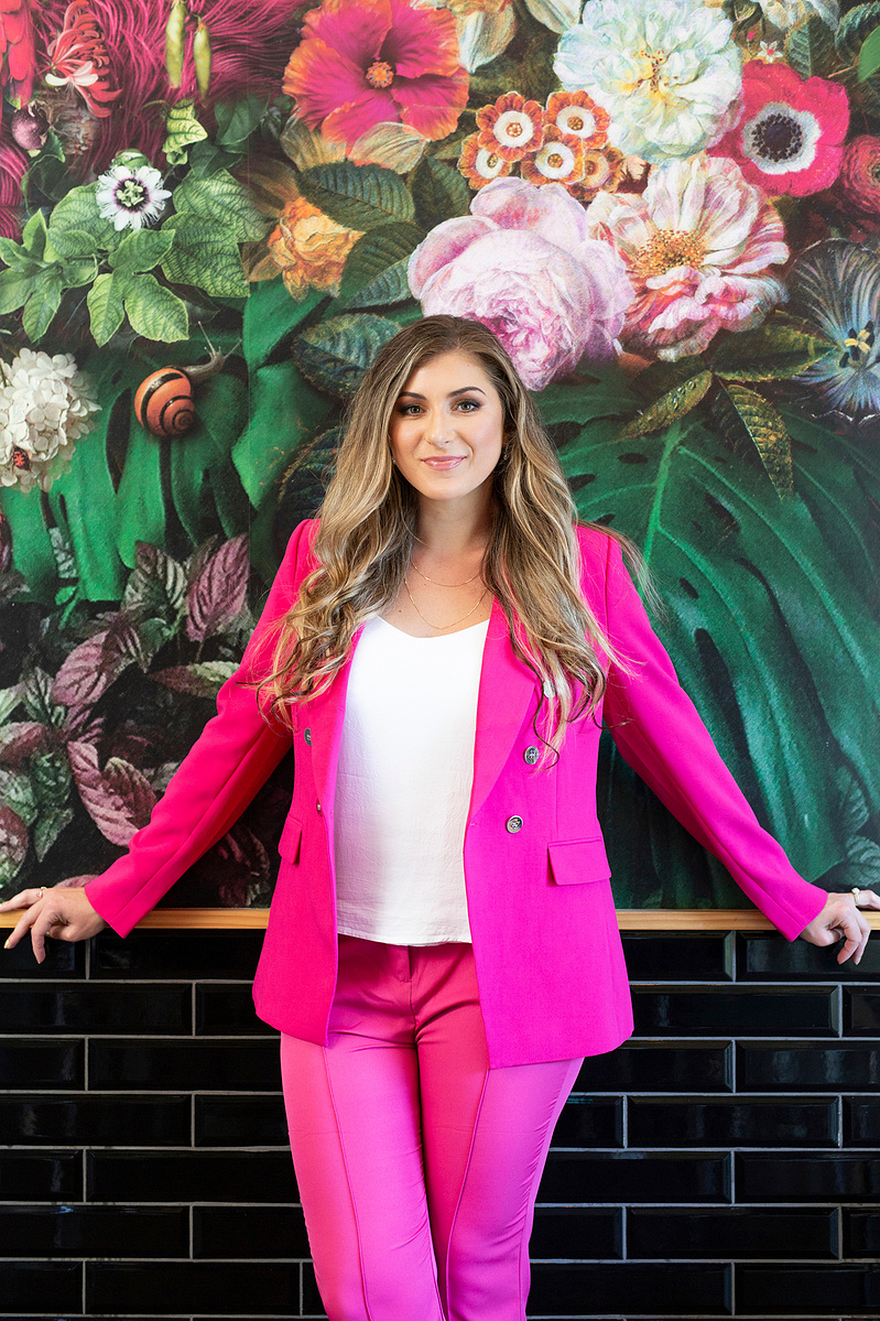 A slim blonde young woman wears a bright pink suit as she leans against a wall covered in floral wallpaper
