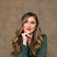 A young woman with long curled hair sits with her chin resting on a hand, she smiles enigmatically off camera