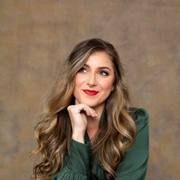 A young woman with long curled hair sits with her chin resting on a hand, she smiles enigmatically off camera