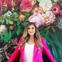 A slim blonde young woman wears a bright pink suit as she leans against a wall covered in floral wallpaper