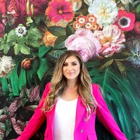 A slim blonde young woman wears a bright pink suit as she leans against a wall covered in floral wallpaper