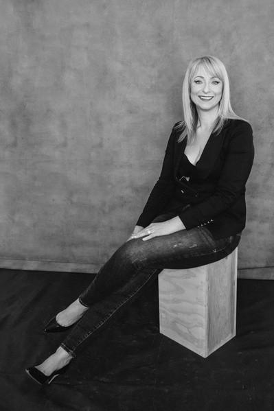A lithe older blonde woman sits on a stool with legs crossed