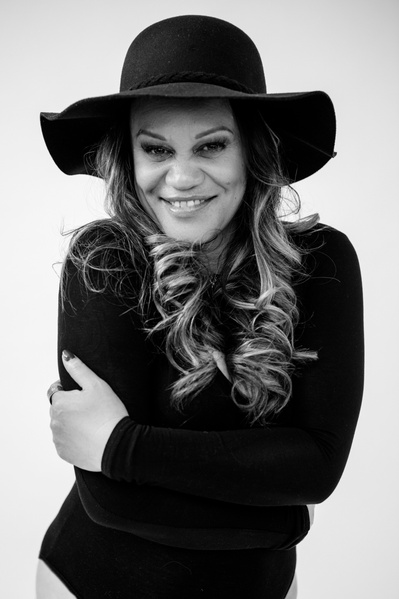 A glamourous woman wearing a black bodysuit and wide brimmed hat embraces herself and faces the camera with a smile