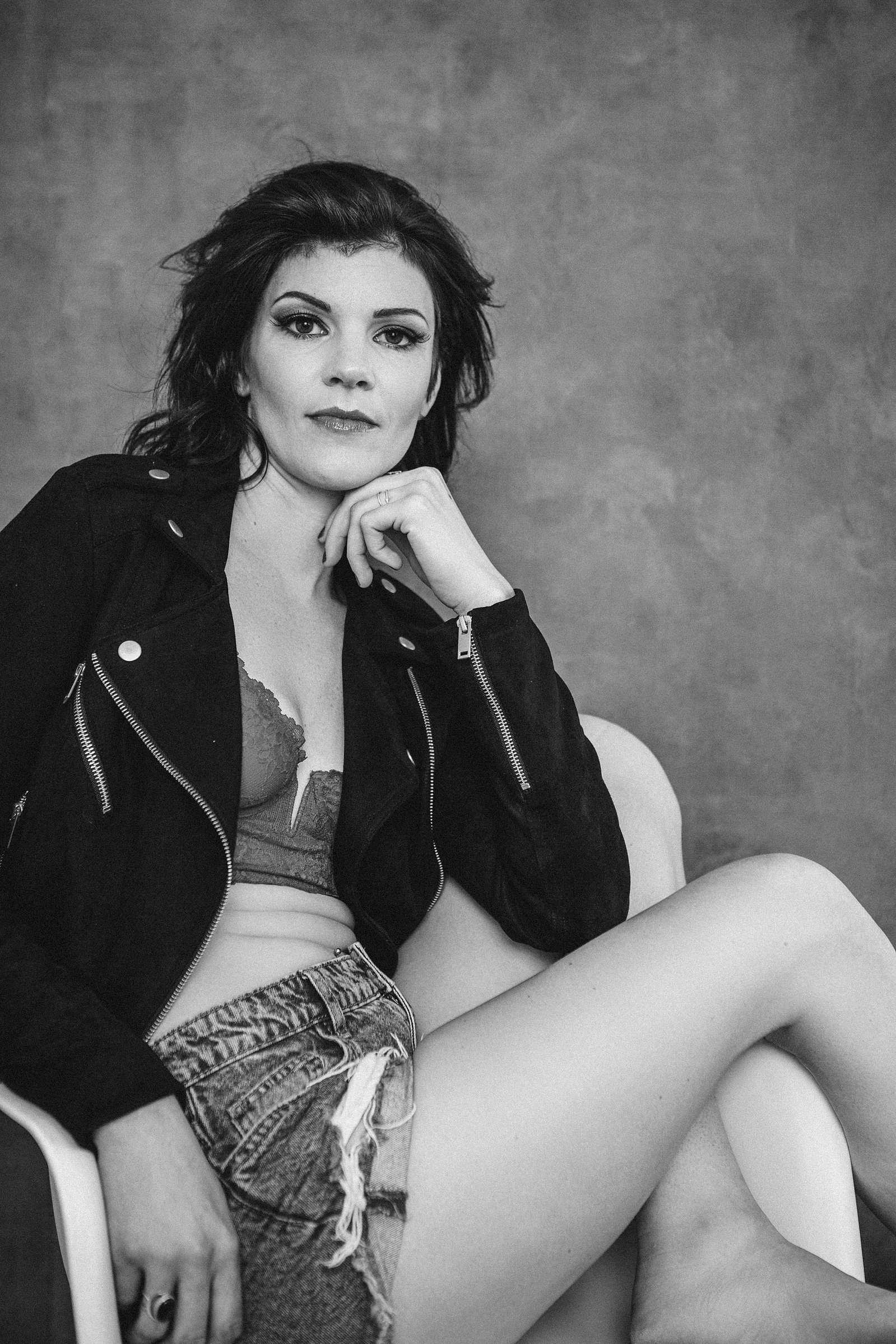 A woman reclines in a white chair, she wears a bralette and ripped denim shorts with a leather jacket on top