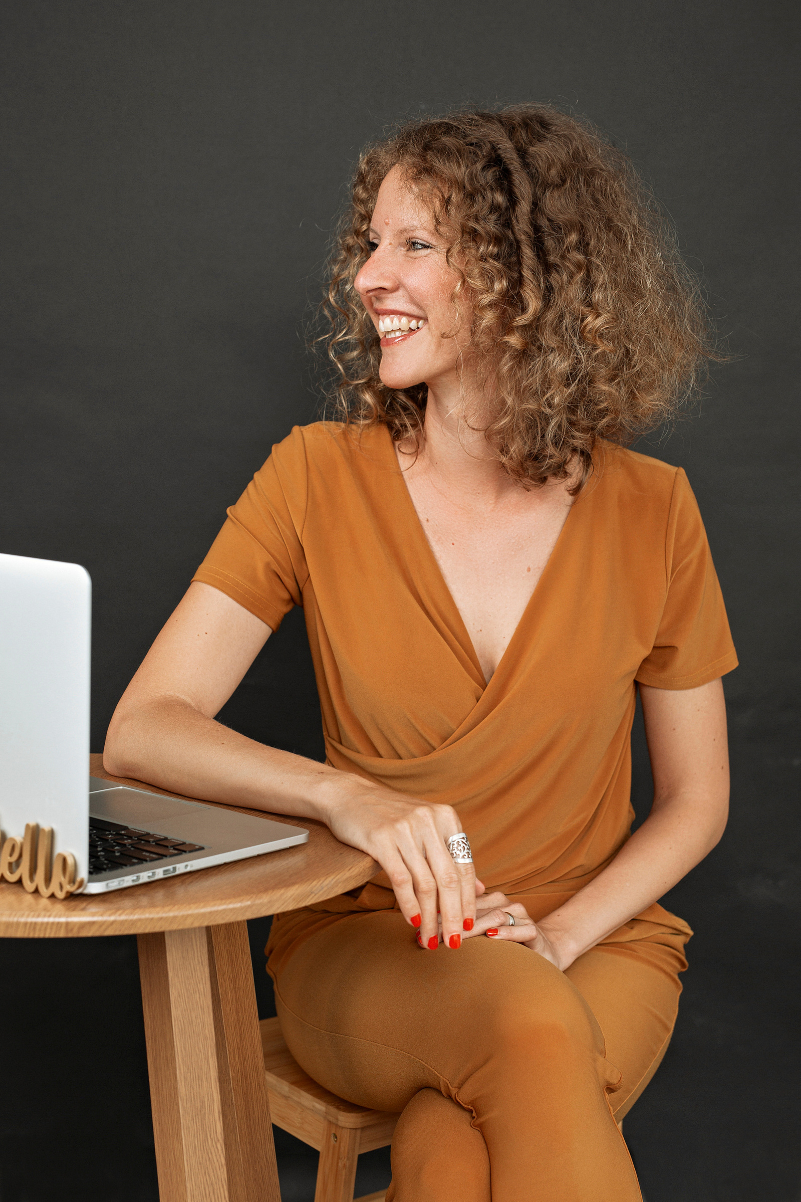 A slim woman with curly reddish hair sits at a desk and smiles with her macbook open