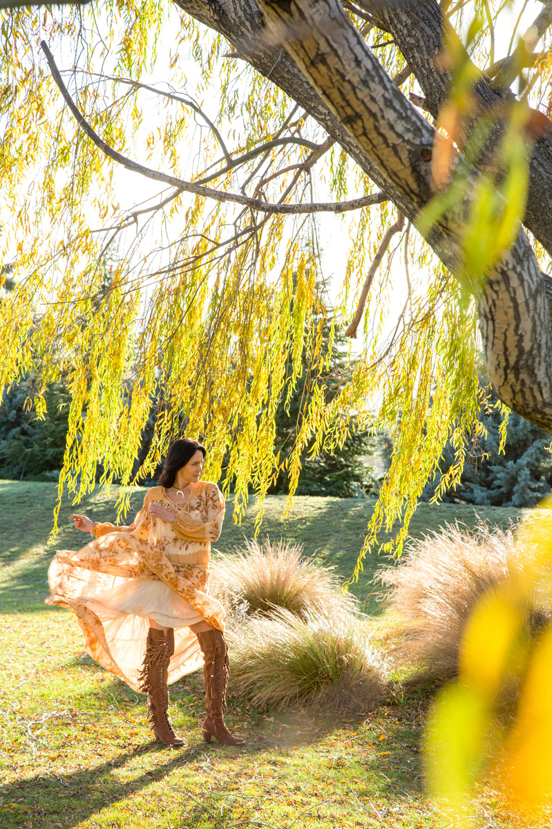 A brunette woman wears a floral dress which she is moving the skirt of, she stands beneath a willow tree and the frame is filled with golden light
