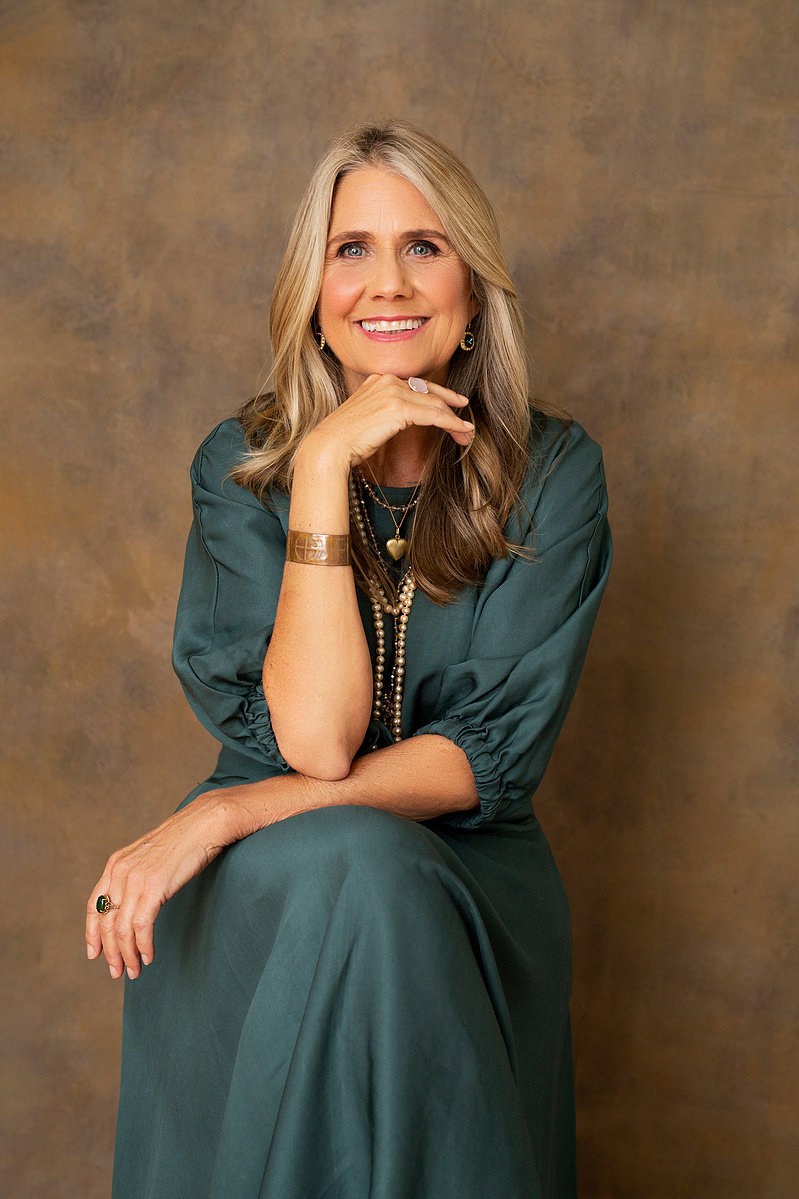 An older woman wearing a green dress smiles into the camera, her chin resting on the back of one hand