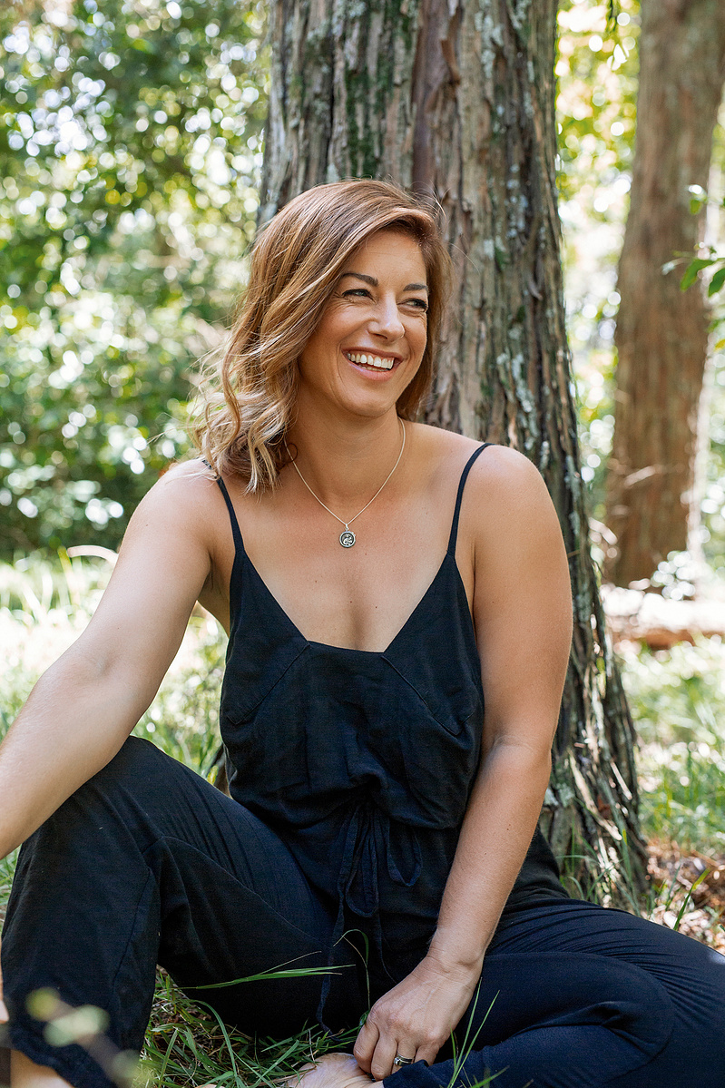 A slim yoga instructor sits under a tree on a sunny day, she is laughing at something off camera