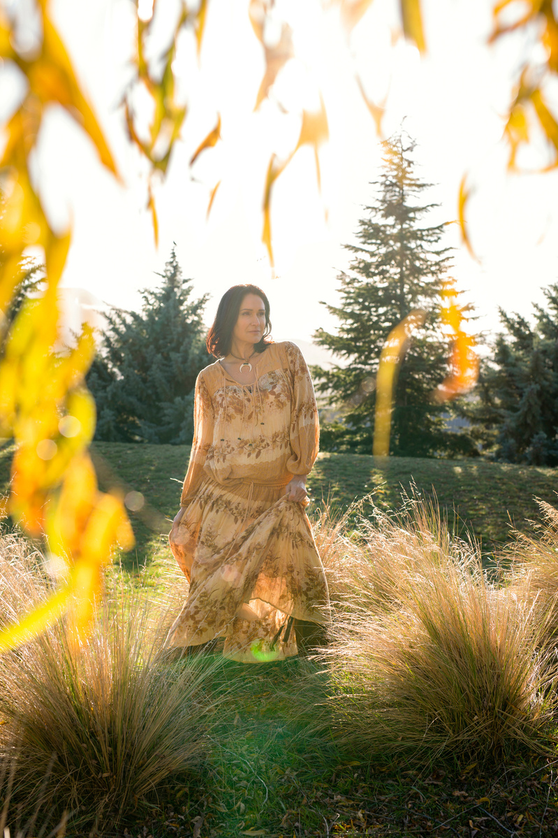 A brunette woman is framed by the branches of a willow tree, she wears a floral dress