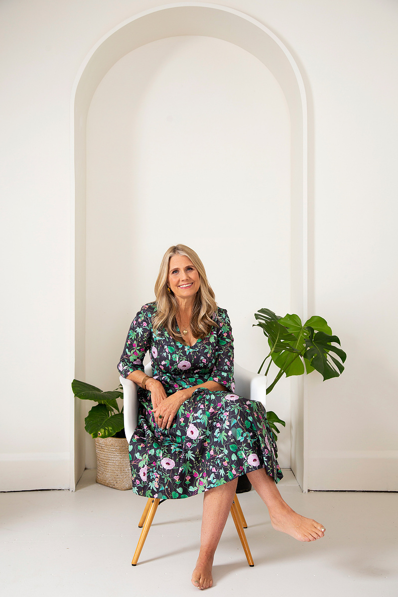An older blonde woman wears a floral dress and sits in a white chair, smiling at the camera