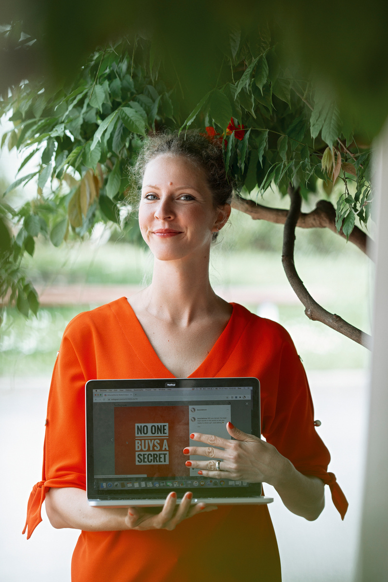 A woman in an orange dress stands under a tree as she proudly displays her laptop screen to the photographer