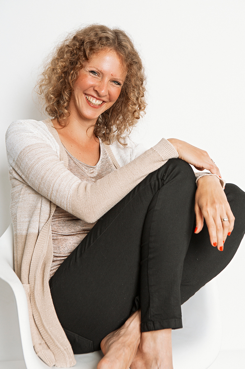 A slim woman with red curly hair sits on a white chair and smiles directly into the camera