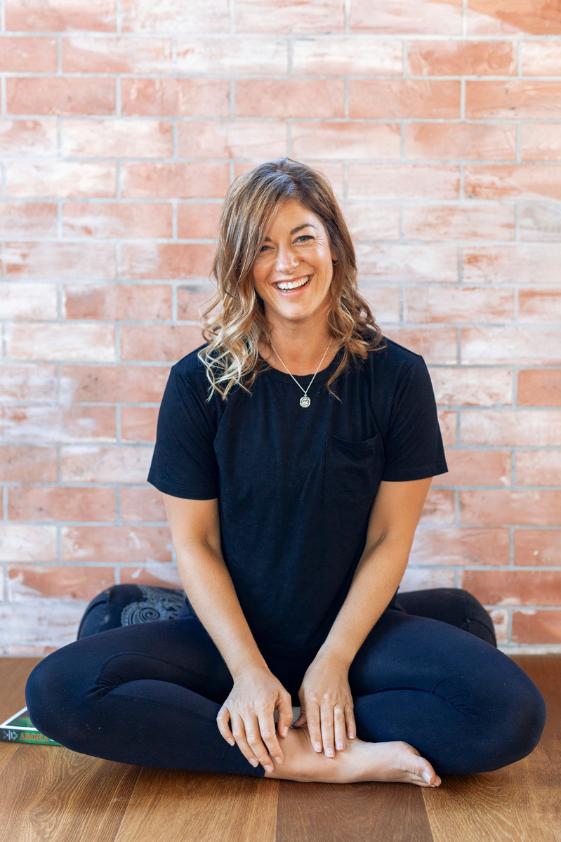 A female yoga teacher dressed all in black sits on a bolster in front of a brick wall