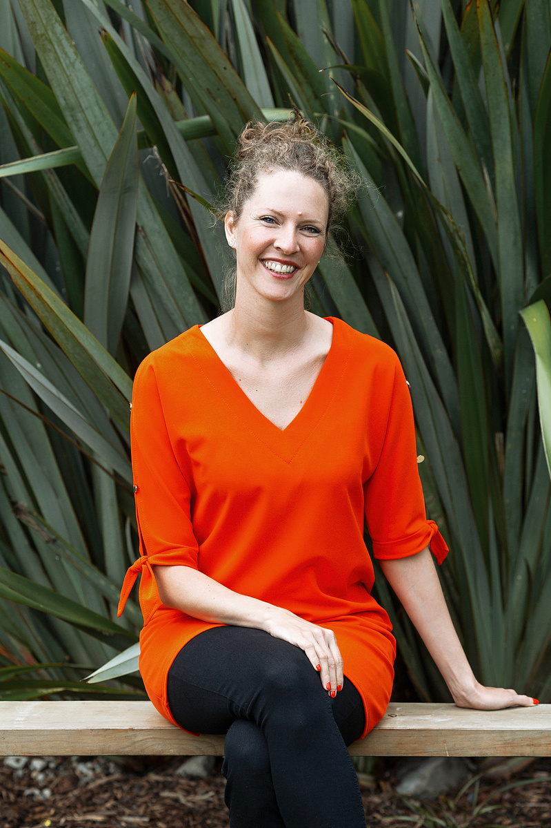 A woman sits on a wooden bench in front of some flax. She has tied up her curly hair and wears an orange dress.